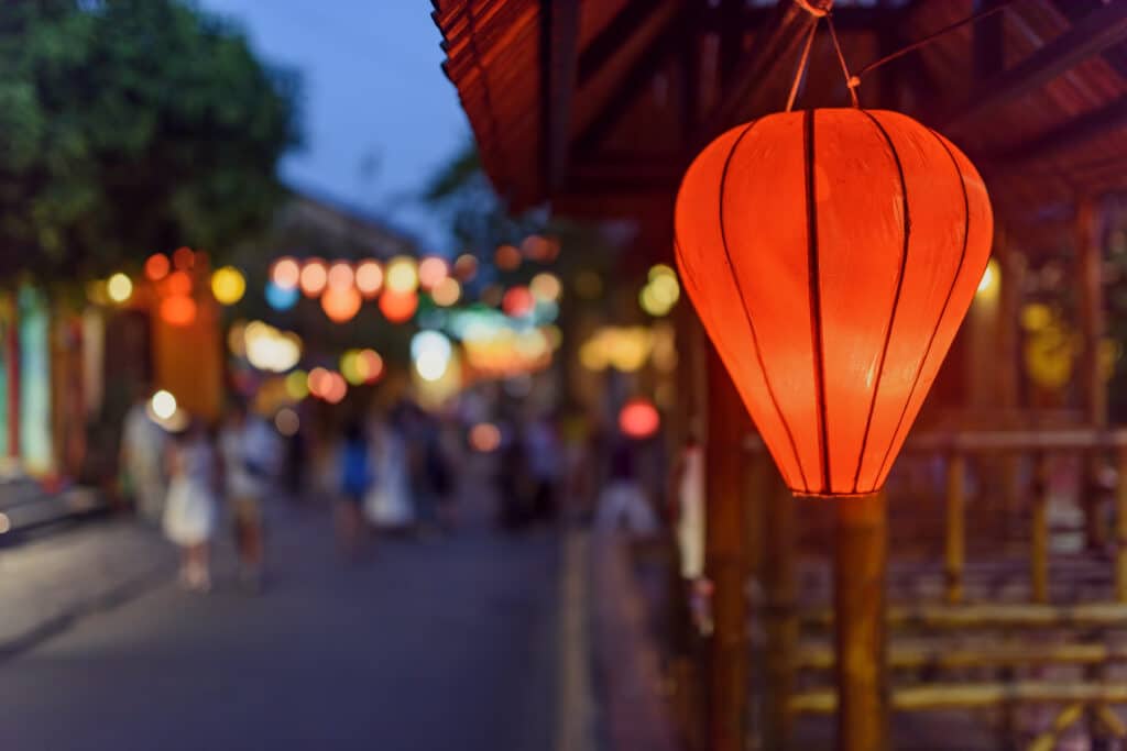 Beleuchtete rote Seide-Laterne in der Altstadt von Hoi An in Vietnam zur blauen Stunde, im Hintergrund schemenhafte Passanten in der Gasse