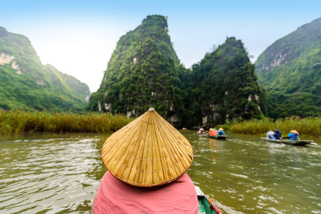Blick aus einem Holzboot auf die Karstlandschaft von Ninh Bình in Nordvietnam, im Vordergrund eine Bootsführerin mit traditionellem Reishut, im Hintergrund weitere Ausflugsboote auf dem Fluss