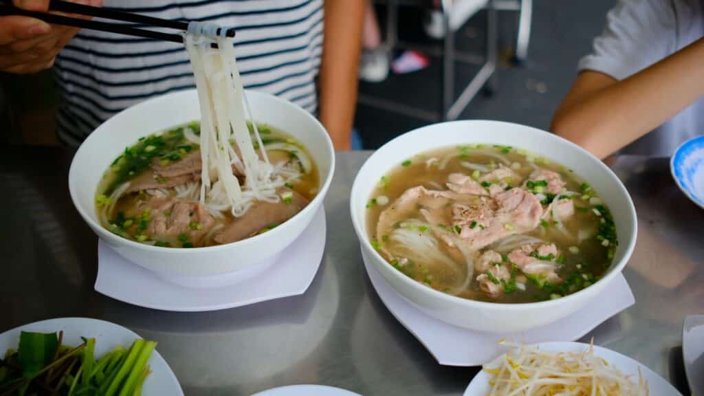 Zwei Schalen vietnamesische Pho-Suppe mit Reisnudeln, Rindfleisch und Frühlingszwiebeln in einer Garküche, Stäbchen heben Nudeln aus der Brühe, daneben ein Teller mit Kräutern und Sojasprossen