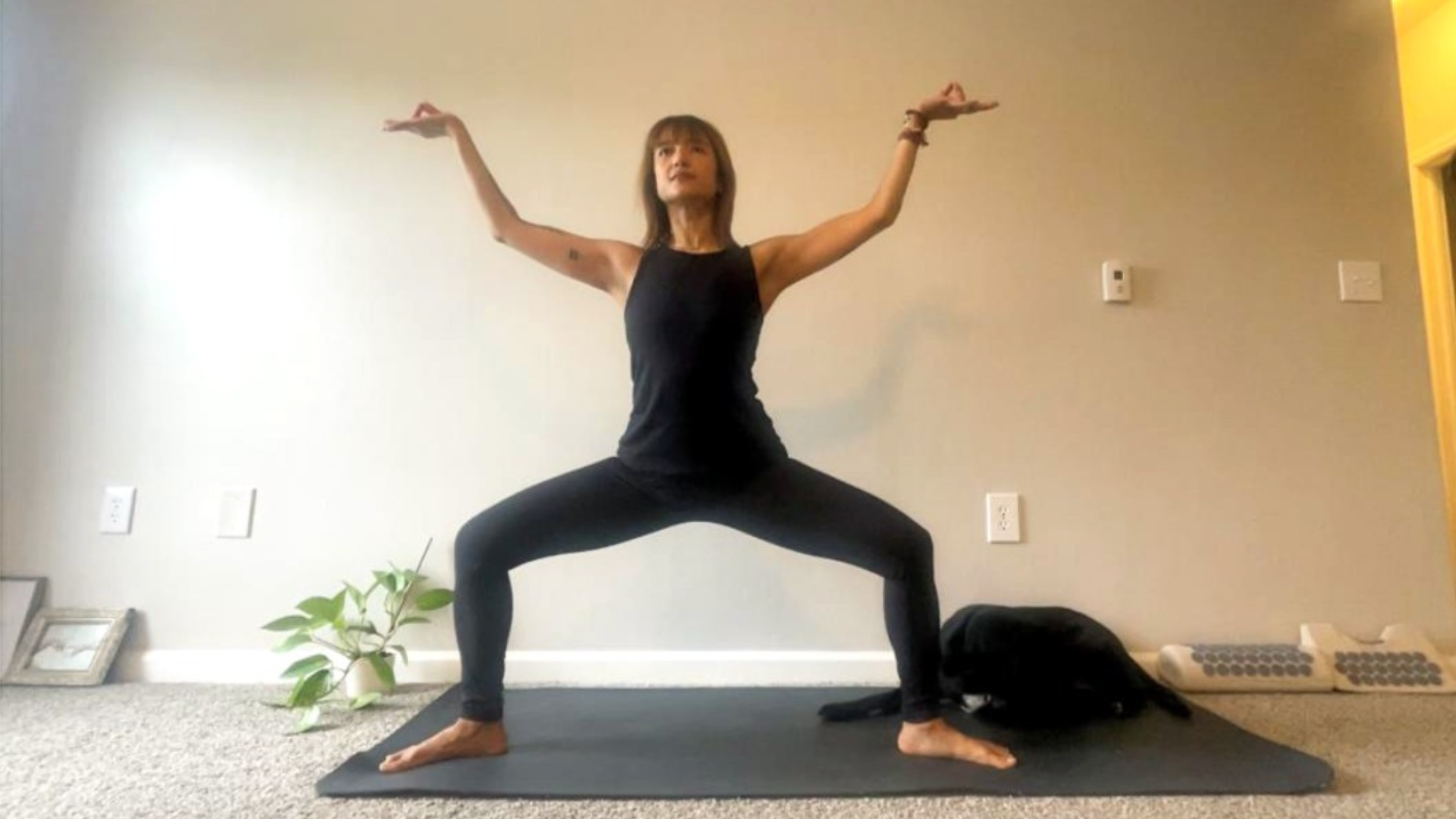 Woman standing on a yoga mat in Goddess Pose while pushing her hands away from her 