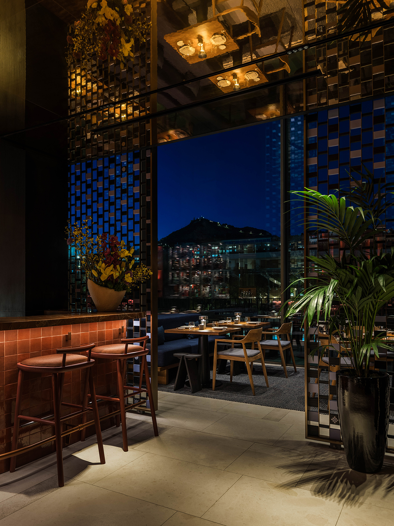 Modern restaurant interior at night with wooden furniture, decorative plants, and a cityscape view through geometric glass panels. Tables are set for dining.