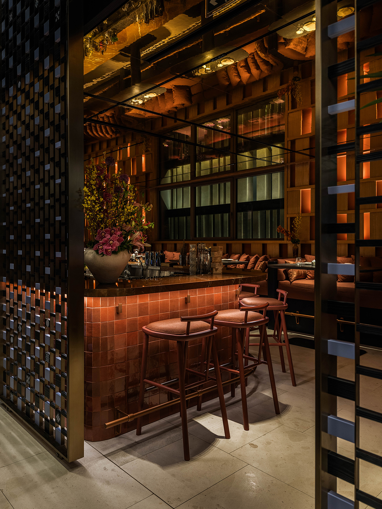 Modern bar area with a tiled counter, wooden stools, floral arrangement, warm ambient lighting, and a decorative metal partition.