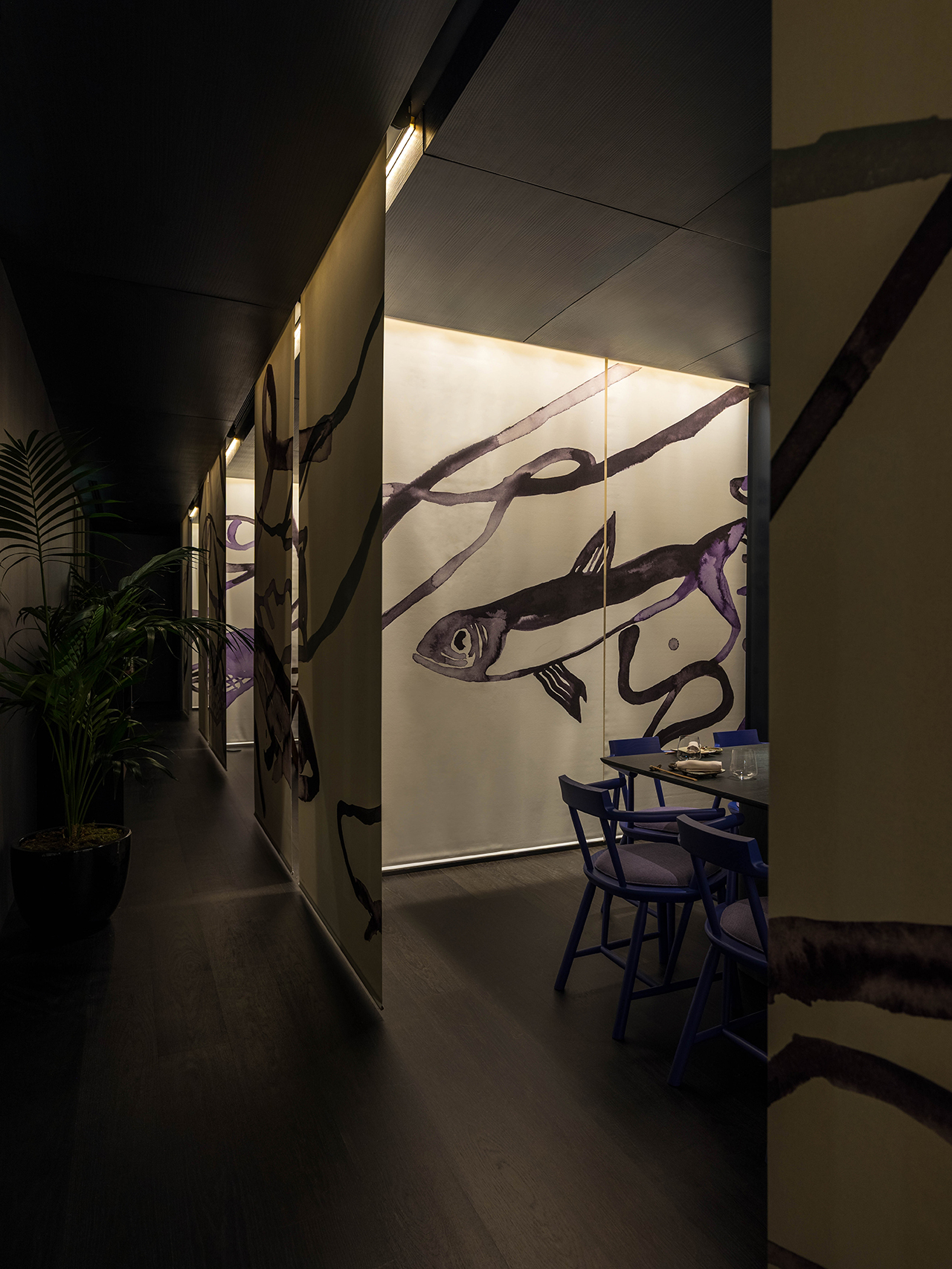 A dimly lit hallway with large wall art featuring a fish and swirling lines, purple chairs around a table, and a potted plant to the side.