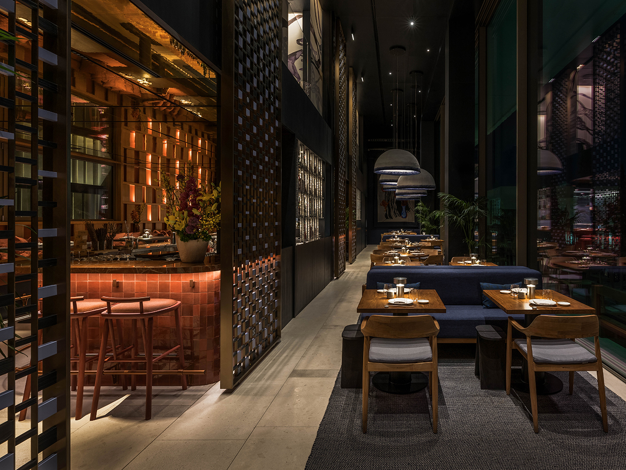Modern restaurant interior with dim lighting, wooden tables set for dining, blue cushioned seating, decorative screens, and large pendant lights.