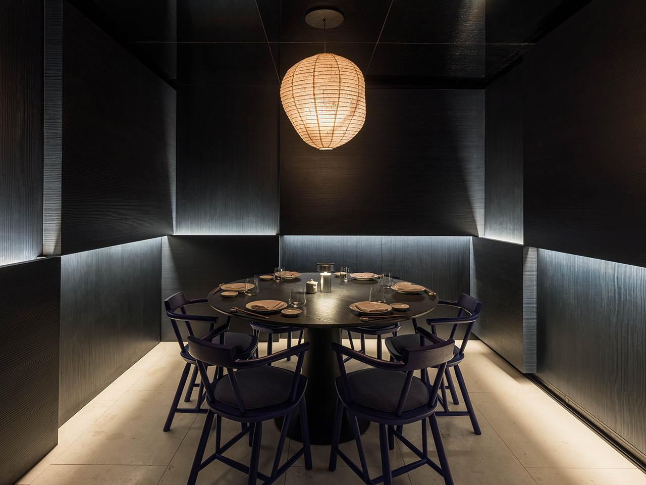 A dimly lit modern dining room with a round black table, seven blue chairs, place settings, and a central hanging woven light fixture.
