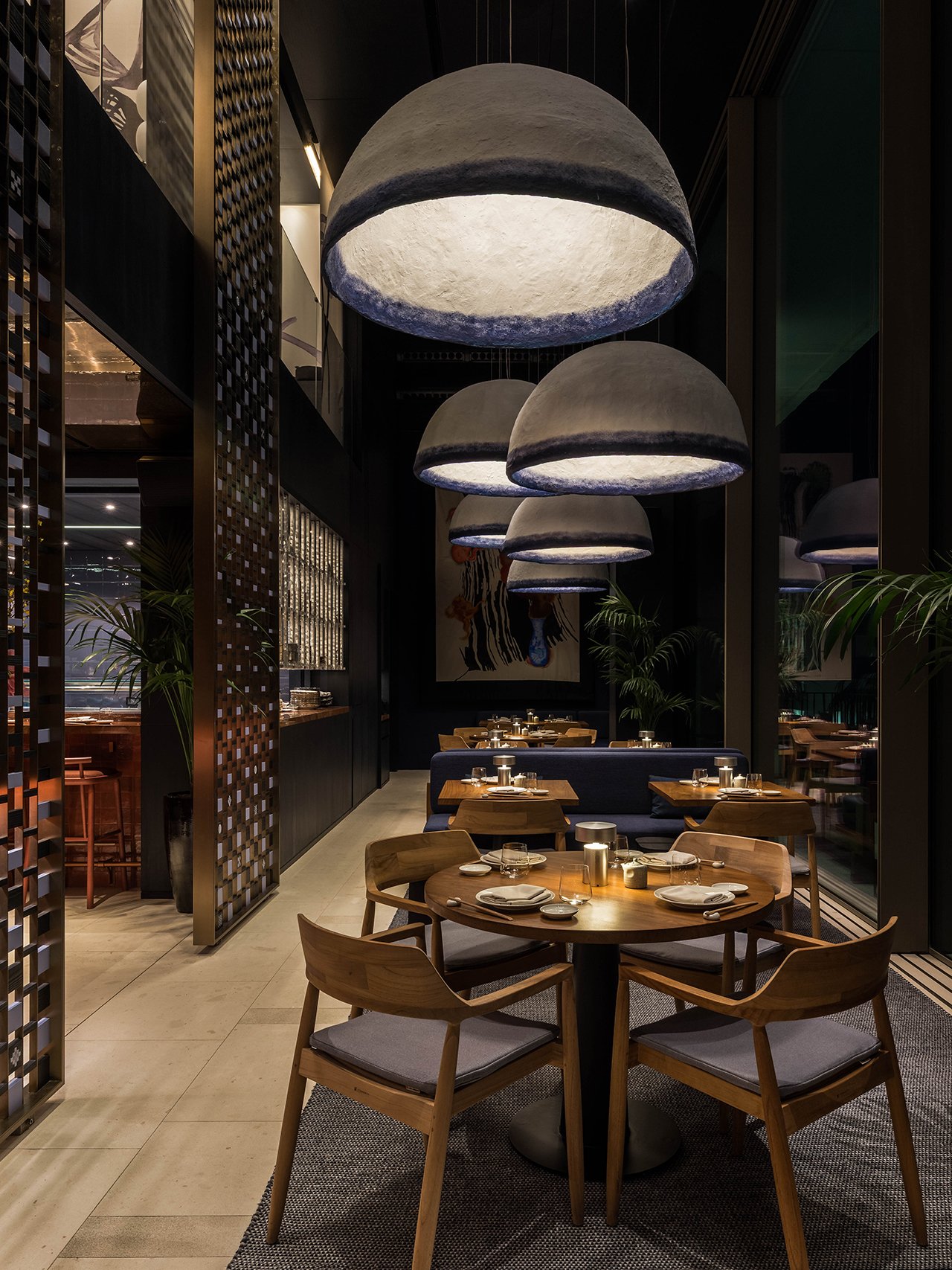 A modern restaurant interior with wooden tables and chairs, low lighting, large dome pendant lights, and place settings arranged for dining.