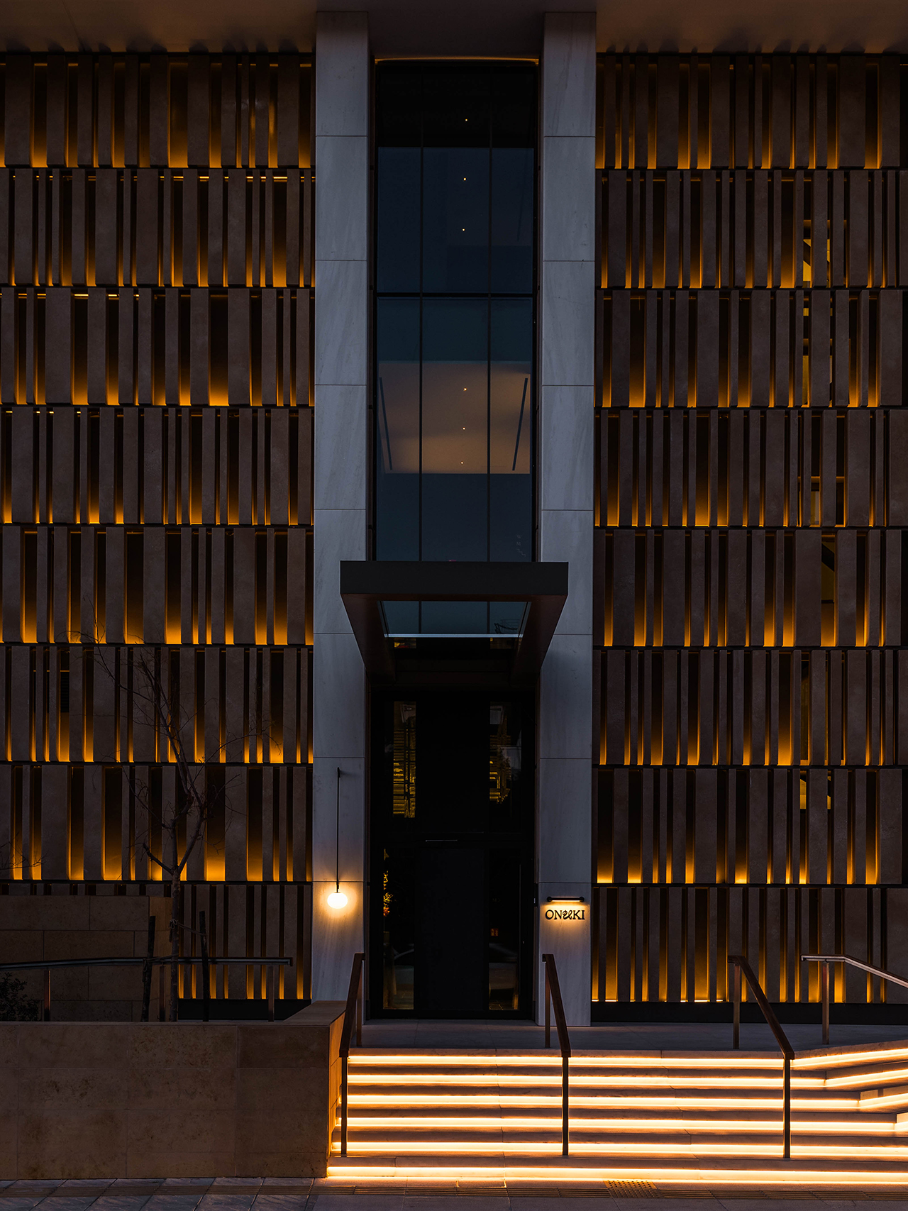 Modern building facade at night with vertical wooden slats illuminated by warm lights, a central glass entrance, and glowing steps leading to the main door.