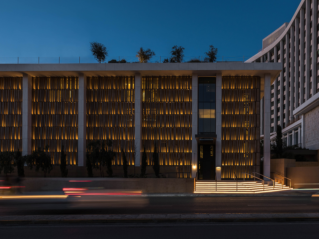 Modern building with vertical slats illuminated by warm lights at night, with blurred car lights passing by and trees visible on the roof.