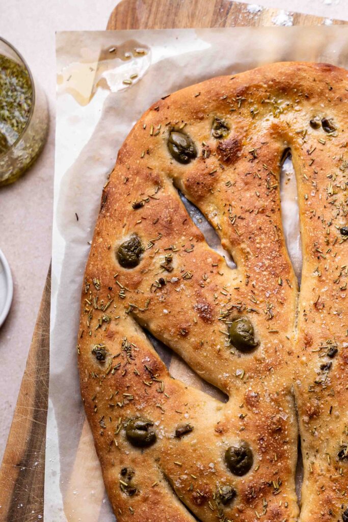 Freshly baked fougasse bread with olives and herbs is placed on parchment paper on a wooden surface, with a jar of oil nearby.