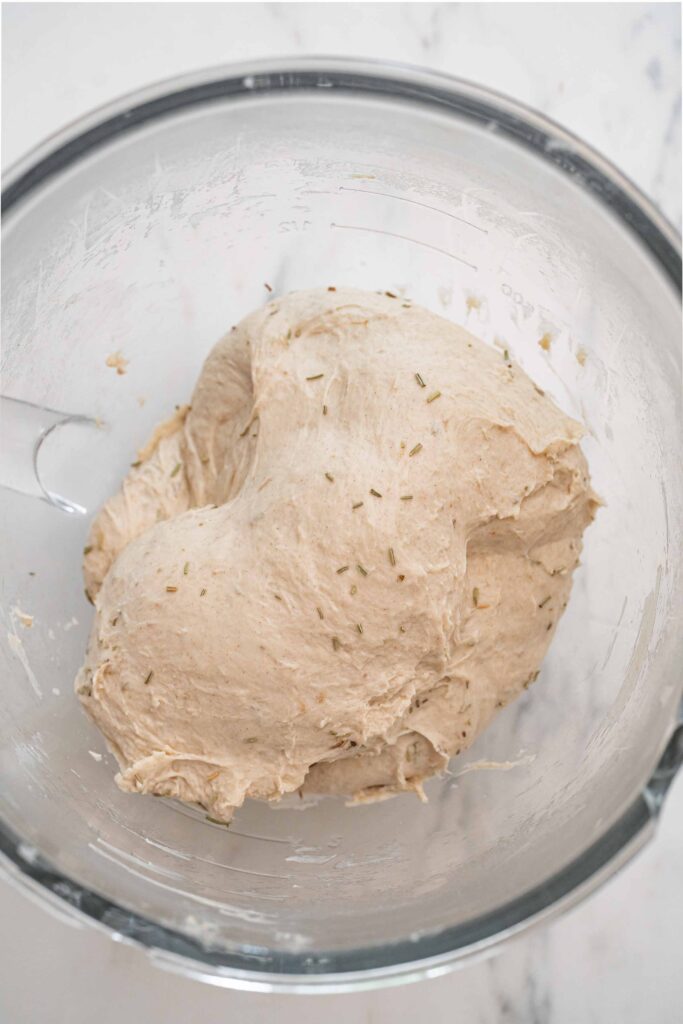 A glass bowl containing leavened bread dough mixed with visible herbs, placed on a light-colored surface.