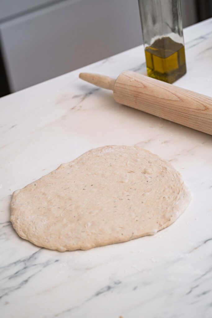 A rolled dough on a marble countertop, with a wooden rolling pin and a bottle of olive oil nearby.