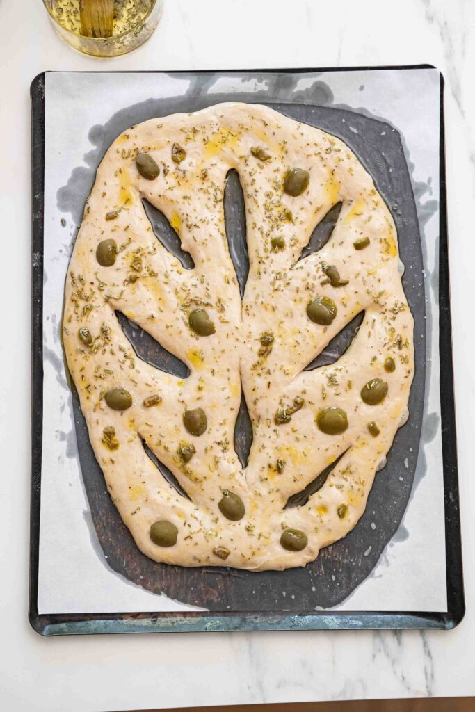 A leaf-shaped fougasse loaf, garnished with green olives and herbs, is placed on parchment paper on a baking sheet.