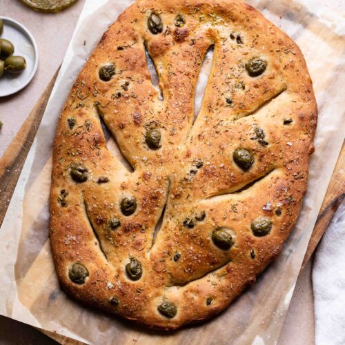 Un pain fougasse fraîchement cuit avec des olives vertes et des herbes sur du papier sulfurisé posé sur une planche en bois, avec un pinceau à pâtisserie et un bol d'huile d'olive à proximité.