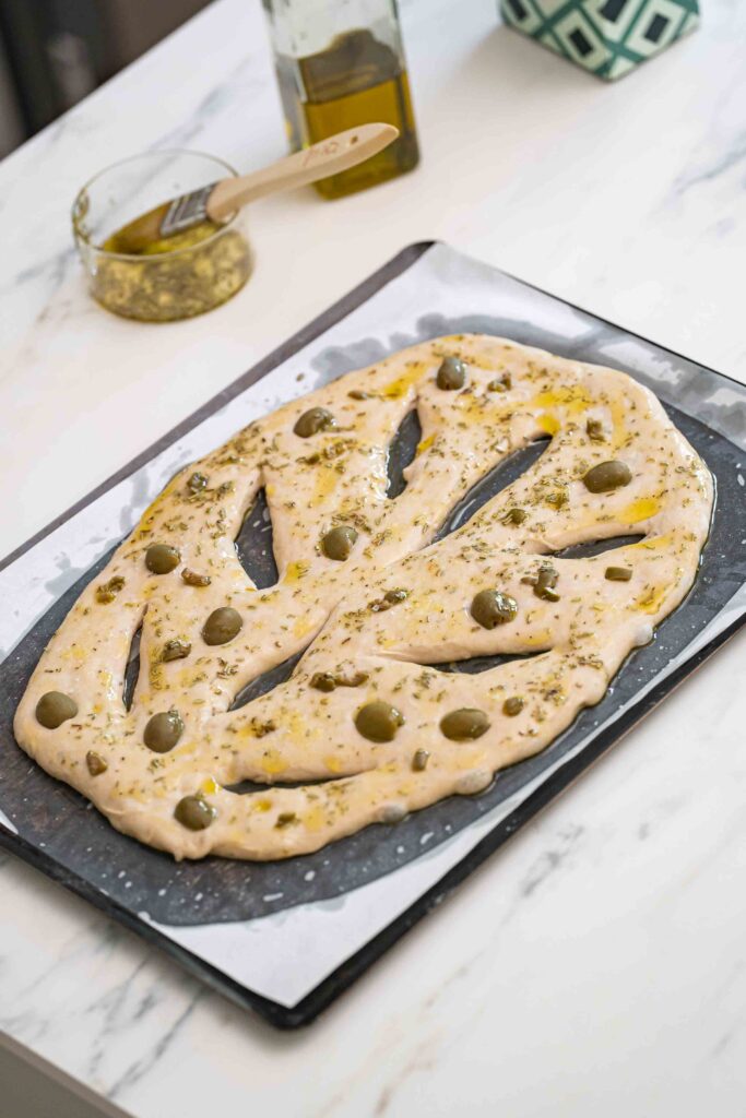 Unbaked fougasse bread dough topped with green olives and herbs on a baking sheet, with olive oil and a brush in the background.