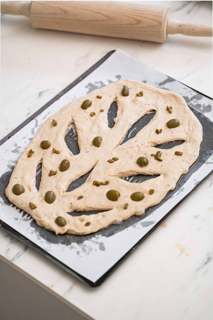 Raw leaf-shaped bread dough with olives and herbs on a parchment-lined baking sheet, with a rolling pin in the background.