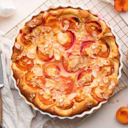 Un clafoutis aux abricots tout juste sorti du four repose sur une grille de refroidissement, parsemé d'amandes effilées et entouré d'abricots frais, d'un couteau, de coquilles d'œufs et d'un petit bol d'amandes.