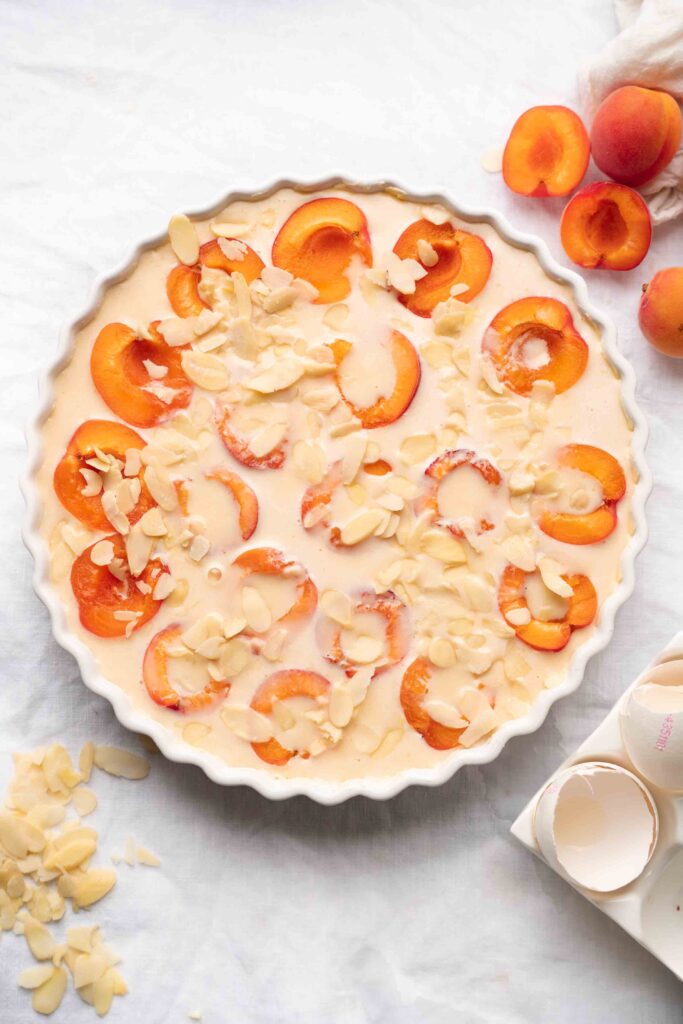 A round baking dish filled with a creamy batter, topped with halved apricots and slivered almonds, sits on a white surface, with slivered almonds and eggshells scattered nearby.