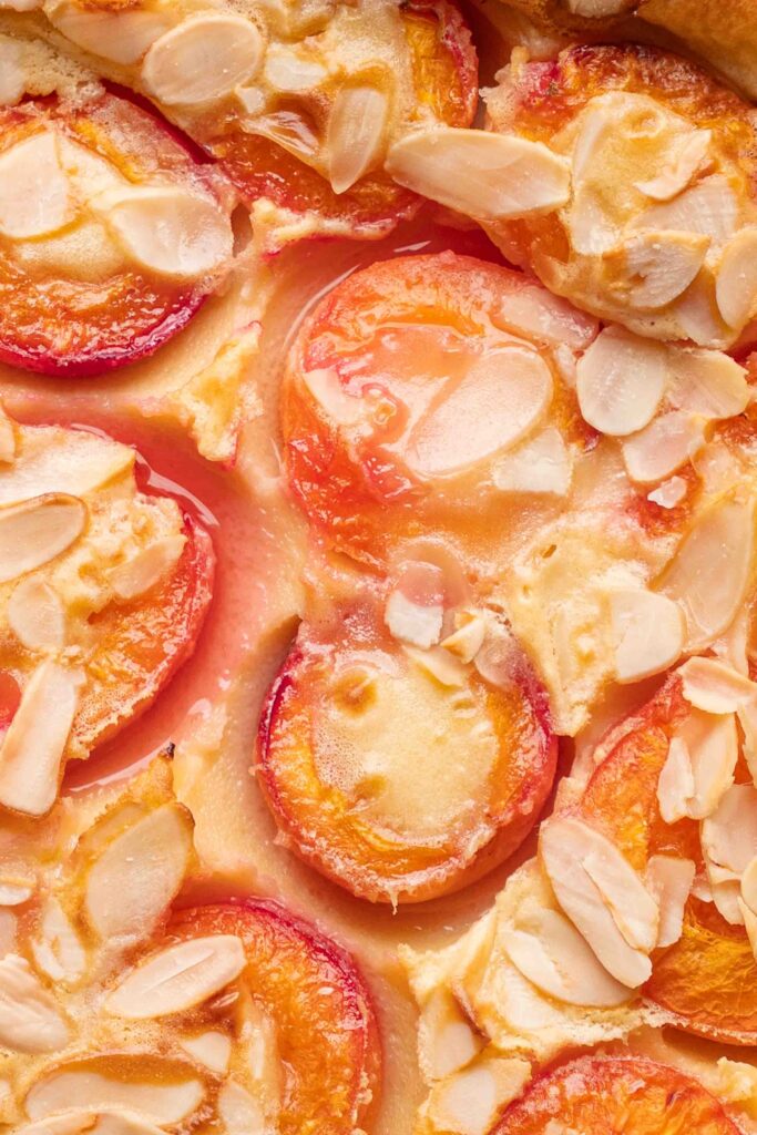 A close-up of a baked tart topped with halved plums and slivered almonds, all glazed with a rich pastry cream.