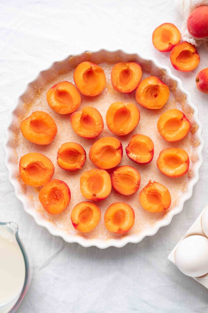 A white tart pan filled with apricots cut in half and arranged, cut-side up, on a bed of pastry, with eggs, whole apricots, and a pitcher of cream placed next to it on a white surface.