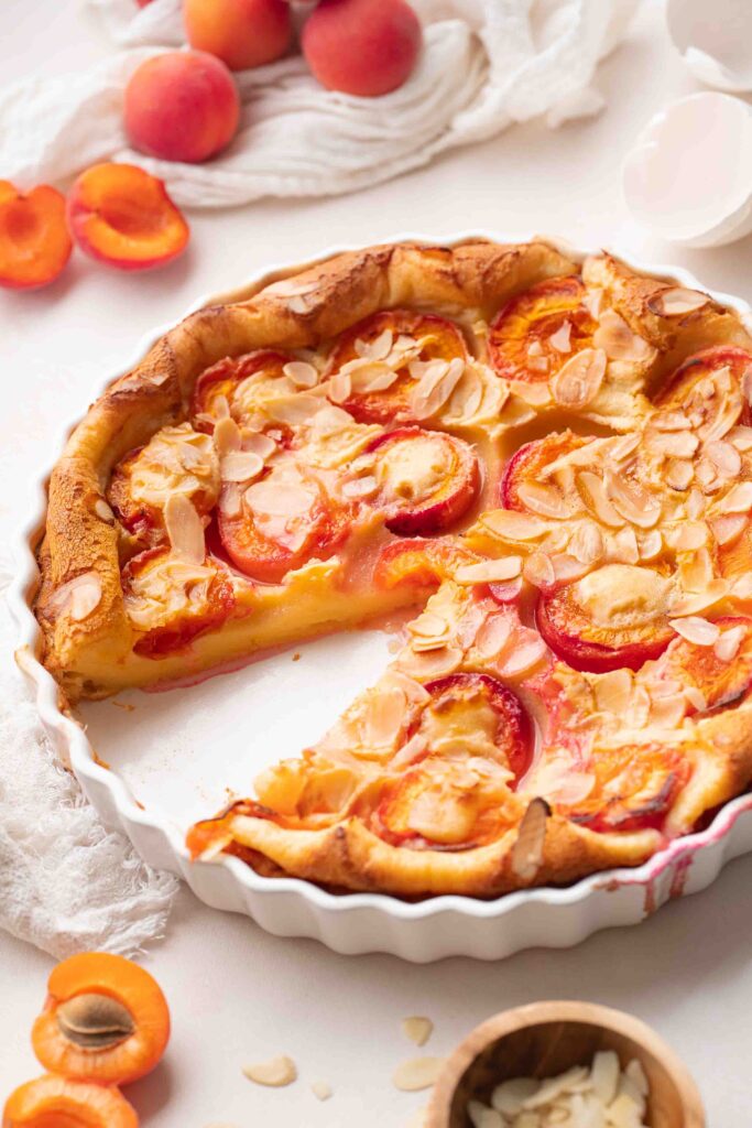 A baked apricot tart, topped with slivered almonds, in a white dish, with one slice removed. Fresh apricots and slivered almonds are arranged on a white surface.