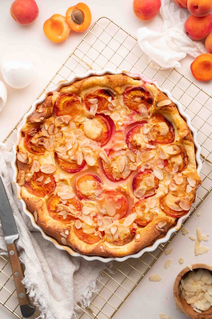 A baked apricot tart, topped with slivered almonds, sits on a cooling rack, surrounded by fresh apricots, a knife, and a linen dish towel.