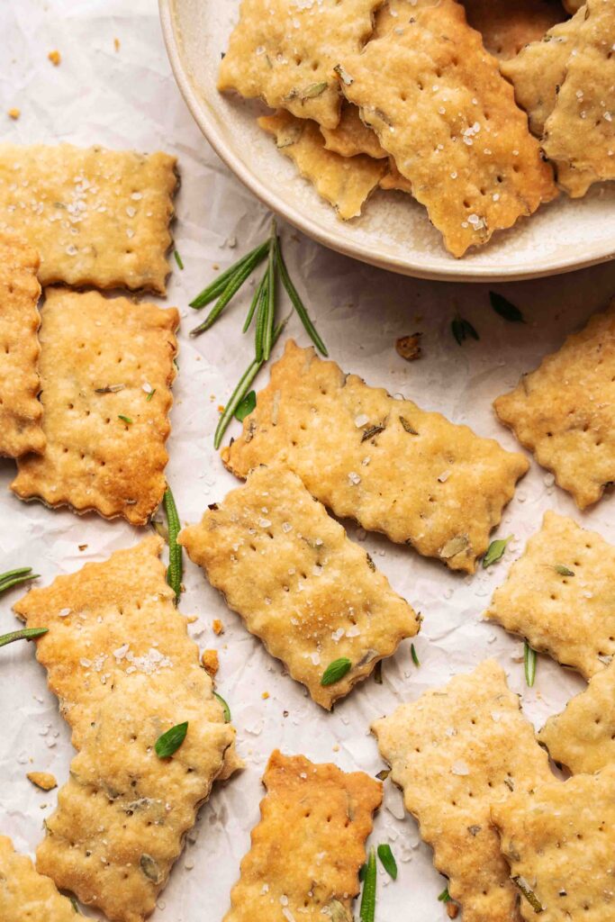 Rectangular crackers with scalloped edges are arranged on a white surface, accompanied by sprigs of rosemary, next to a bowl filled with more crackers.