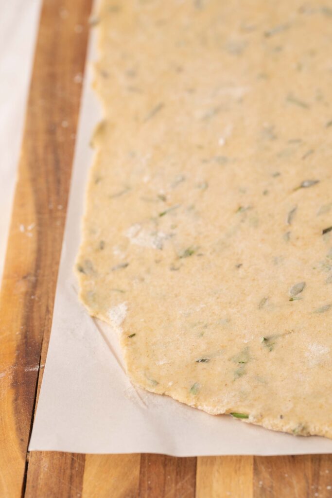A sheet of rolled-out dough, sprinkled with herbs, rests on parchment paper placed on a wooden work surface.