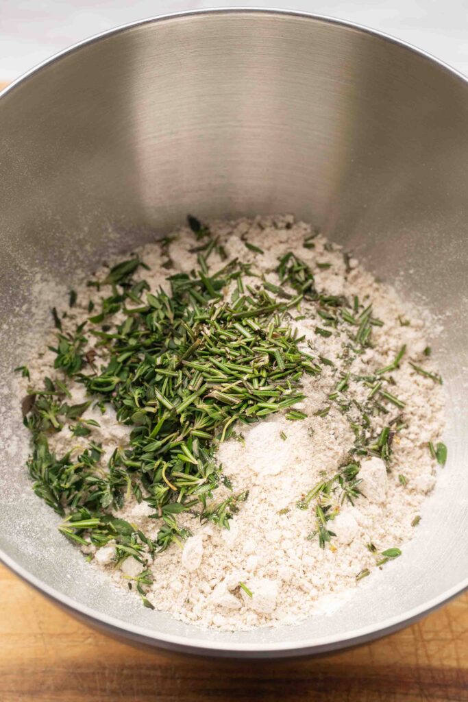 A metal mixing bowl containing flour and chopped fresh herbs, sitting on a wooden countertop.