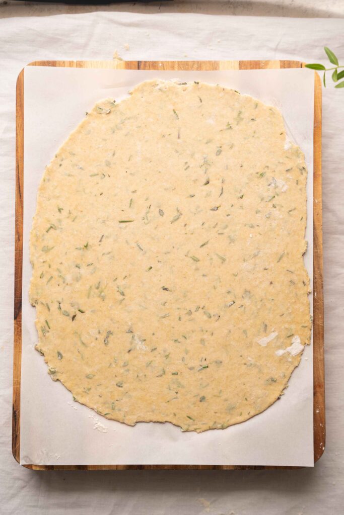 The rolled-out herb dough is placed on parchment paper laid out on a wooden board, ready to be worked with.
