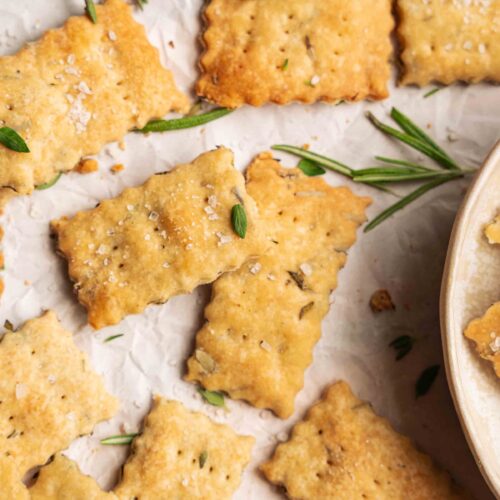 Des crackers maison rectangulaires, salés et aux herbes, sont disposés sur du papier sulfurisé, tandis que d'autres se trouvent dans un bol à droite. On aperçoit également des brins de romarin frais.