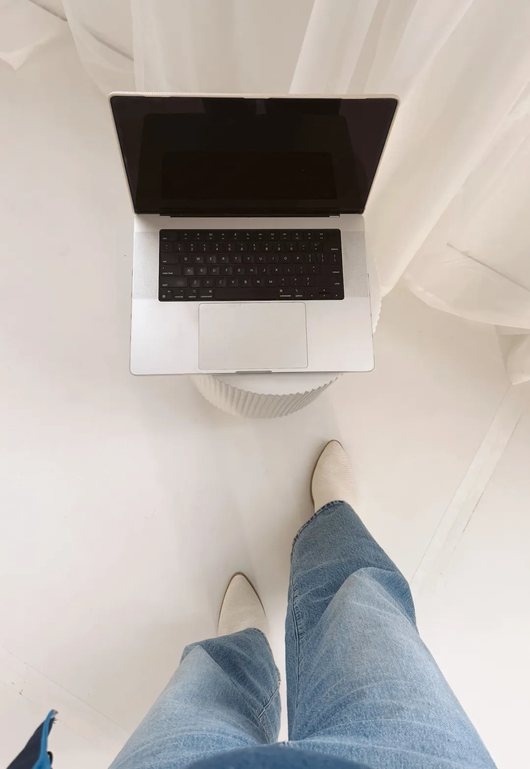 Overhead photo of a work-from-home setup with a silver laptop on a white stool, wide leg jeans, and cream pointed shoes in a bright neutral office space.