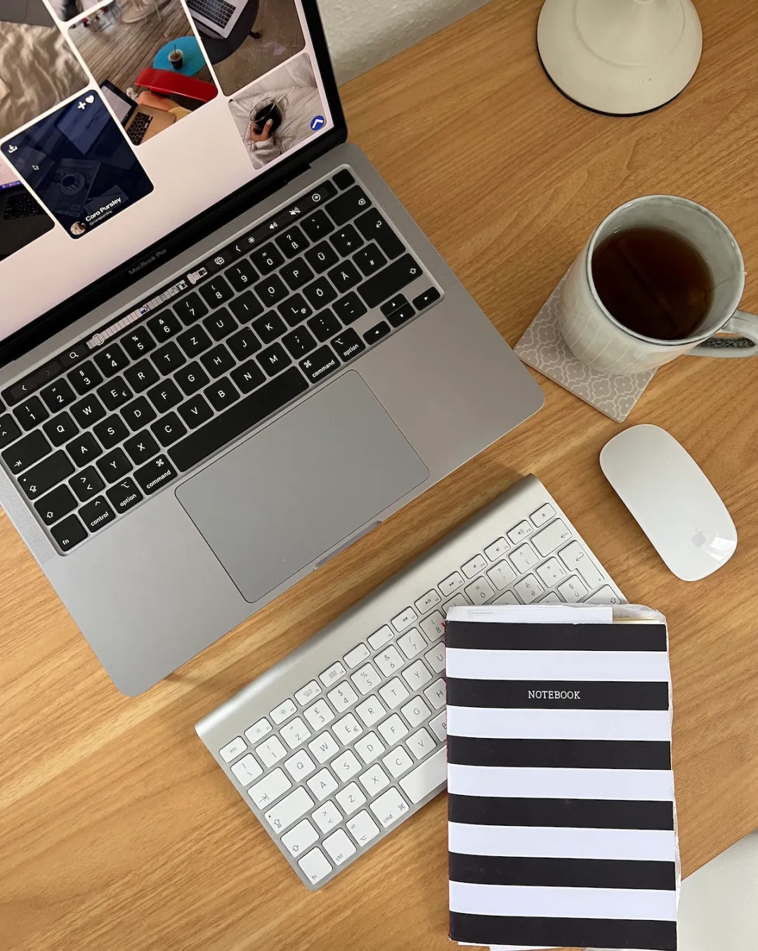 A flat lay of a MacBook Pro open to a Pinterest feed, a striped notebook, an Apple keyboard and mouse, and a cup of tea on a wood desk, representing a business owner locking in on her goals.