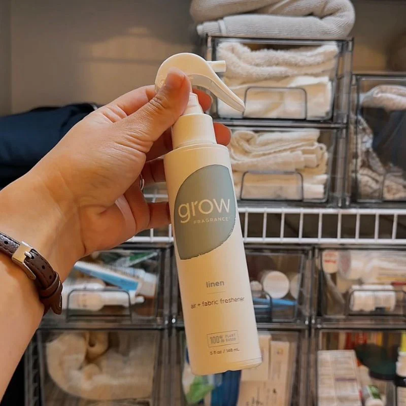 Hand holding a 'grow' linen fabric freshener spray bottle in a linen closet.