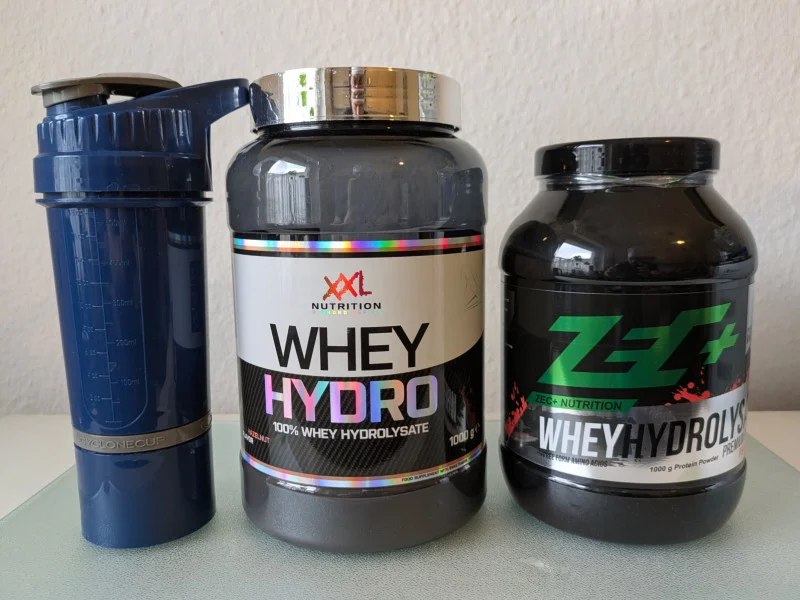Zwei Whey Hydrolysate aus dem Whey Protein Test neben einem blauen Shaker