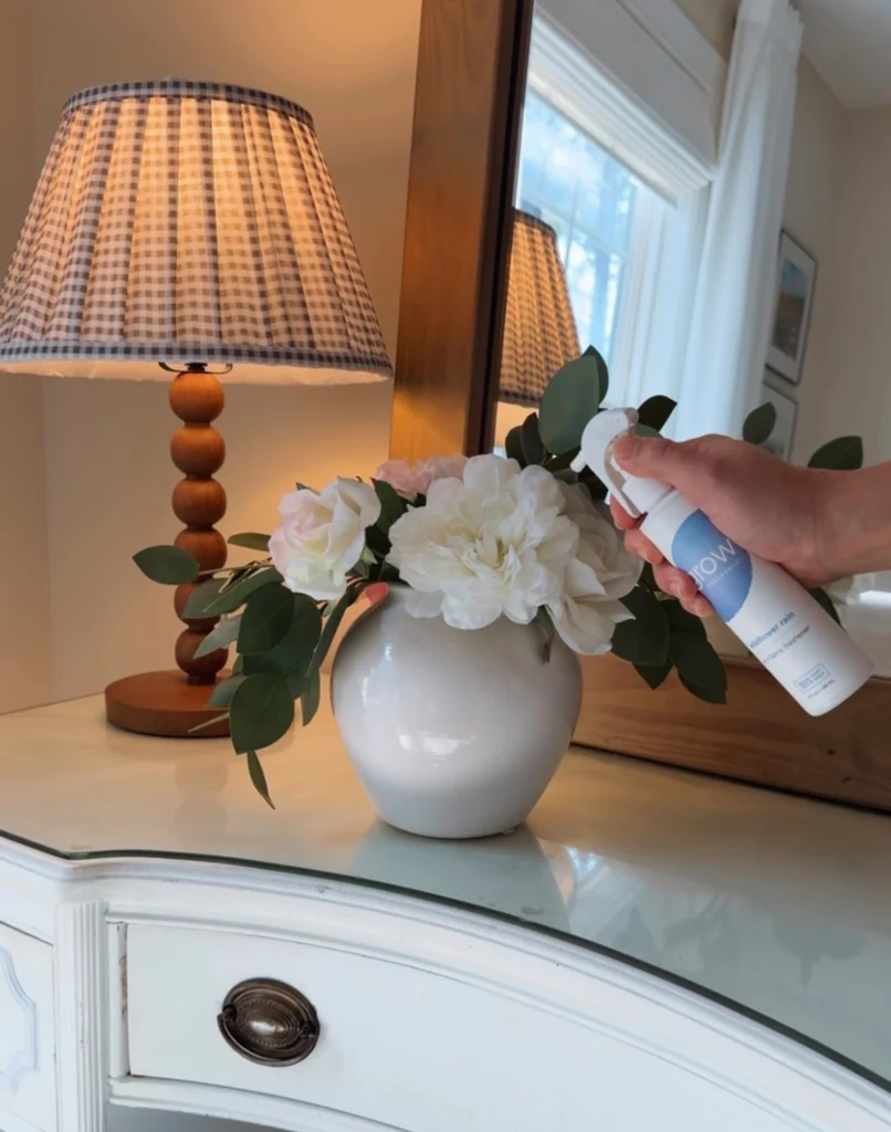 Person spraying a Grow Fragrance air and fabric freshener on a vase with flowers on a dresser next to a lamp
