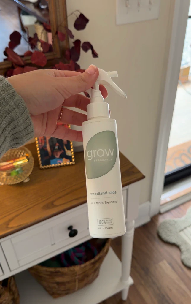 Hand holding a Grow Fragrance air and fabric freshener product bottle with a spray nozzle on a wooden surface.