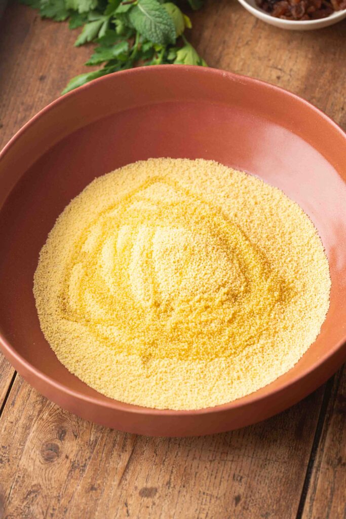 A large brown bowl filled with uncooked yellow couscous sits on a wooden table, with green herbs partially visible in the background.