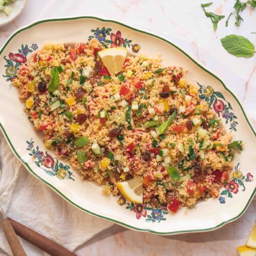 Un plat de salade de couscous au taboulé, composé de tomates coupées en dés, de concombres, de maïs, de raisins secs, d'herbes hachées et de quartiers de citron, est servi dans un plat à motifs floraux, à côté de cuillères de service.