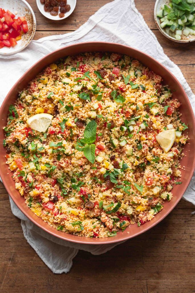 A large bowl of taboulé, a French couscous salad with chopped vegetables, garnished with lemon wedges and mint leaves, on a wooden table. Small bowls of diced tomato, cucumber, and raisins are nearby.