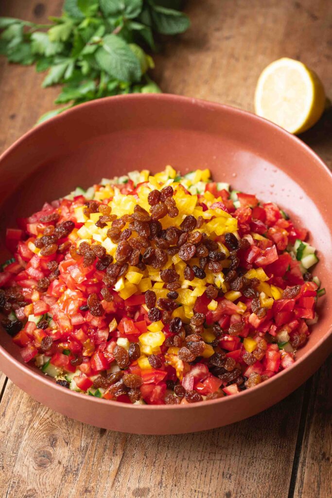 A bowl filled with diced tomatoes, yellow bell peppers, cucumber, and topped with golden raisins, set on a wooden surface with mint and a lemon in the background.