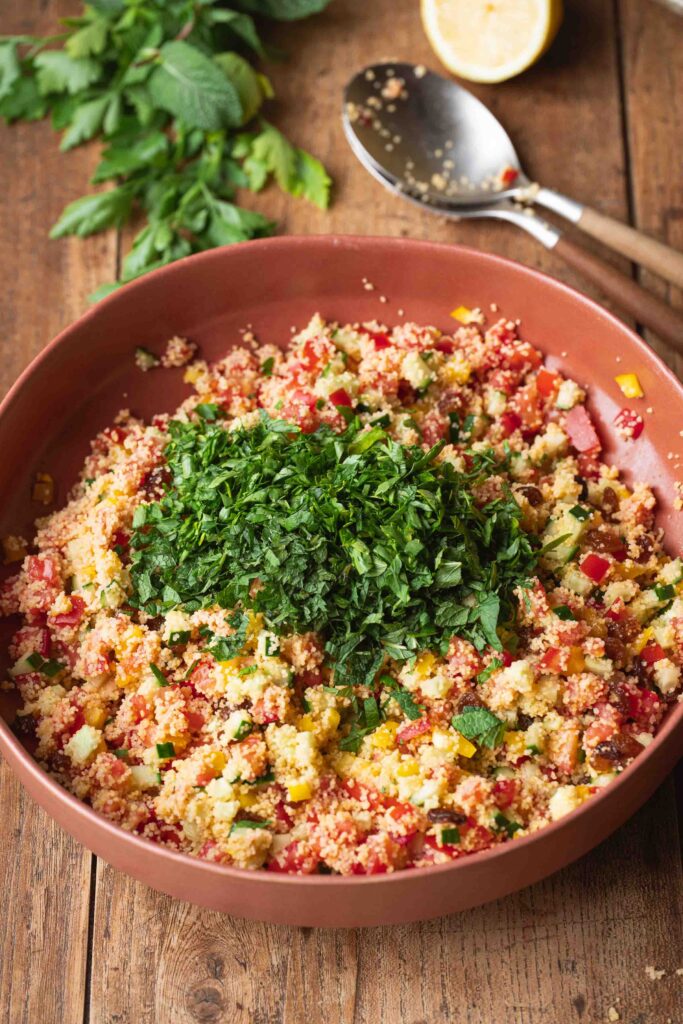 A bowl of taboulé, a French couscous salad with chopped fresh herbs on top, surrounded by a bunch of parsley, a lemon half, and a serving spoon on a wooden table.