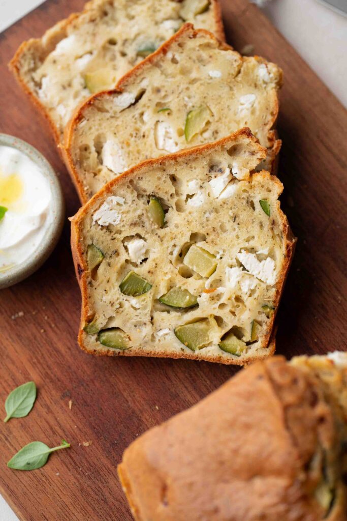 A slice of zucchini bread, with chunks of zucchini and feta clearly visible, resting on a wooden board next to a small bowl of yogurt.