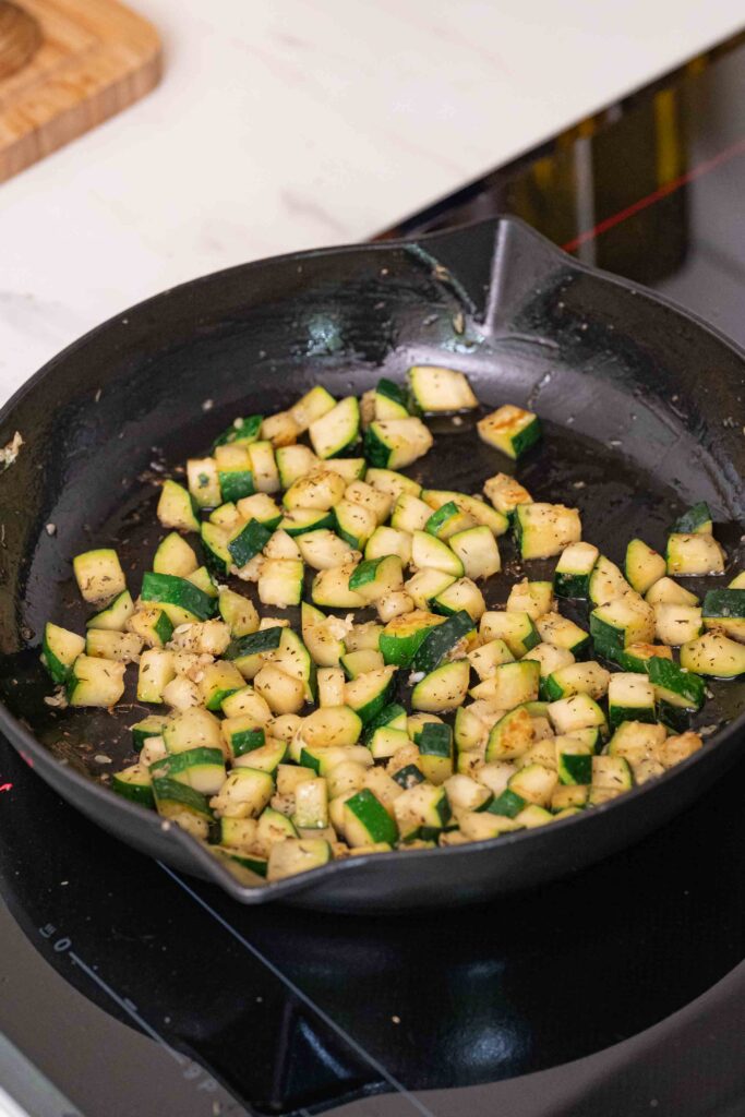 Diced zucchini, sizzling in a black cast-iron skillet on the stove, seasoned with herbs and spices.