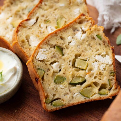Des tranches de pain salé, dans lesquelles on distingue des morceaux de courgette et de fromage, sont disposées sur une planche en bois, à côté d'un petit bol de sauce crémeuse agrémentée d'herbes aromatiques.