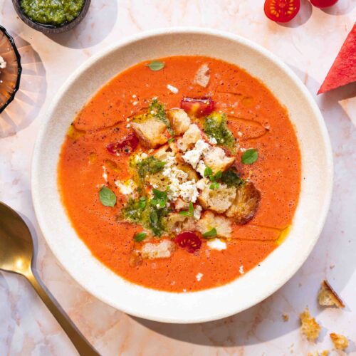 Eine Schüssel Tomaten-Gazpacho, garniert mit Croutons, Feta, Pesto und Kräutern, umgeben von Wassermelonenscheiben, Kirschtomaten und einem Löffel.