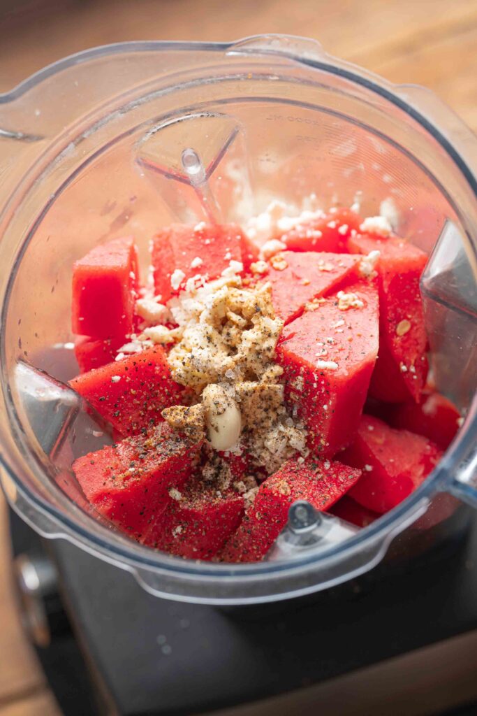 Pieces of watermelon, a clove of garlic, ground black pepper, and crumbled cheese in the blender bowl, ready to be blended.
