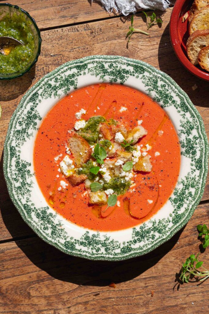 A bowl of tomato soup topped with croutons, cheese, pesto, fresh herbs, and a drizzle of olive oil, sitting on a wooden table.