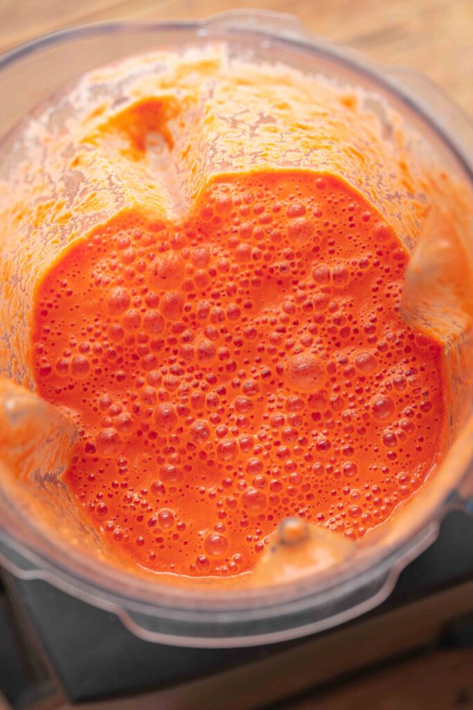 A bright orange watermelon gazpacho being blended in the transparent bowl of a blender, viewed from above, with a texture resembling that of a smoothie or a sauce.