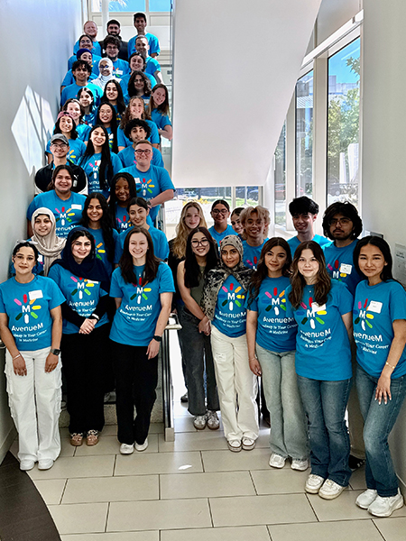 Dozens of community college students in blue "AvenueM" shirts stand in the hallway and a stairwell of a medical school building