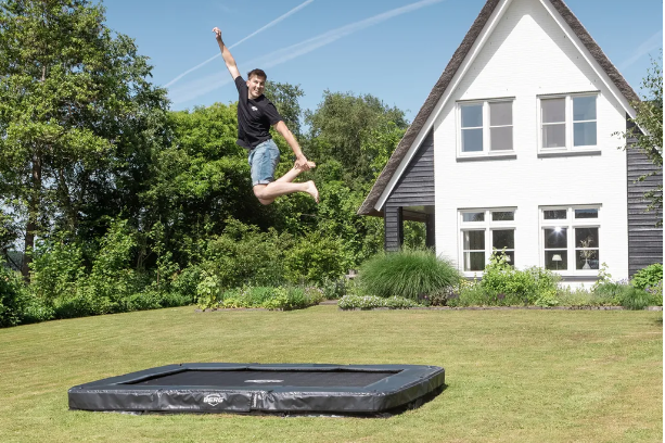 Ein Jugendlicher springt auf einem Berg Trampolin Ultim Champion InGround Sports AirFlow Pro in die Luft. Dabei hat er seinen rechten Arm in die Luft gestreckt, mit dem anderen greift er einen Fuß. Das Trampolin ist in der Erde eingelassen, im Hintergrund ist ein Haus zu sehen, was umwachsen von Bäumen und Sträuchern ist. Der Himmel ist blau.